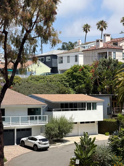 The image shows a street scene in Newport Beach, California, USA, lined with residential buildings. The sky is partly cloudy. Palm trees are visible in the background. The buildings range in style and size, with a mix of white, light green, and gray colors. Some buildings feature red tile roofs. Lush greenery, including trees and hedges, fills the spaces between the houses. A white SUV is parked on the street in front of one of the buildings. A large tree with exposed branches is visible in the foreground. The street is paved and appears to be made of asphalt and brick.