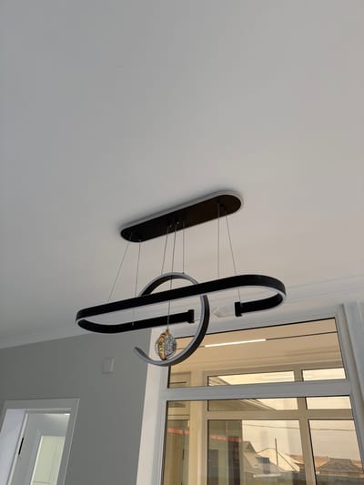 The image shows a modern, black oval-shaped chandelier hanging from a white ceiling. The chandelier has a sleek design with integrated LED lighting. A decorative spherical element, possibly made of glass and metal, is suspended from the center of the chandelier. The chandelier is suspended by thin wires. Below the chandelier, there is a large window with multiple panes, reflecting the interior of the room and the exterior view. The interior appears to be a room with light gray walls. A white door frame is visible to the left of the window. The location is stated to be in the city of Baş-Bulak, Kyrgyzstan.