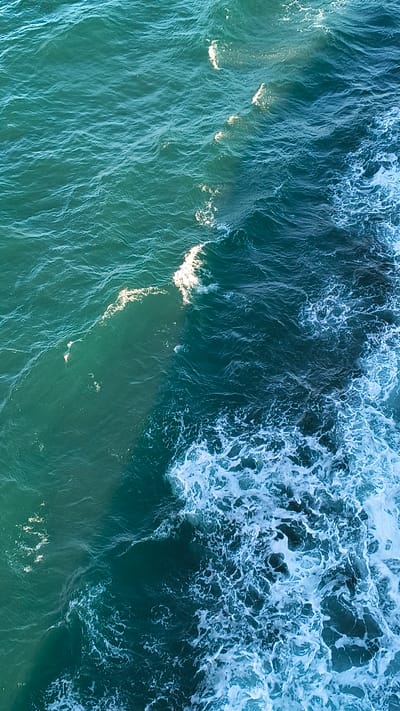 This is an aerial, high-angle, full shot of the ocean. The water is a deep teal color, with lighter turquoise hues where the sunlight hits it. White foam and waves are visible, especially on the right side of the frame, indicating movement and a slightly choppy sea. The texture of the water varies from smooth ripples to churning foam. The scene is devoid of any man-made objects or living beings, presenting a purely natural environment. The location is specified as Unknown, United Kingdom.