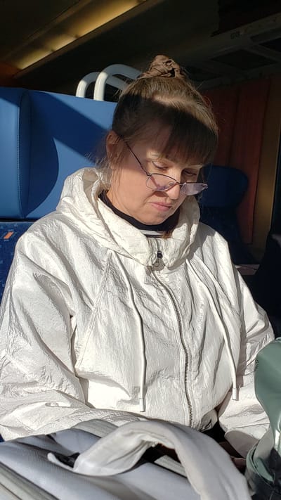 A woman with her hair in a bun is wearing glasses and a white hooded jacket. She is seated on a blue upholstered seat, possibly on a train or bus. The scene appears to be illuminated by natural light, casting shadows on her face and clothing. In the background, there are white arched structures and a red curtain, suggesting an indoor environment. The overall impression is one of a passenger resting or looking down. The location is identified as Važec, Slovakia.