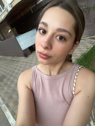 A young woman with brown hair and brown eyes is taking a selfie outdoors. She is wearing a pink ribbed tank top with a white and gold chain strap on her left shoulder. Her left arm is visible, and she has a mole on her inner forearm. Behind her, a sign on a building reads "РЕМОНТ ОБУВИ ИЗГОТОВЛЕНИЕ КЛЮЧЕЙ," which translates to "SHOE REPAIR KEY MAKING." The ground is paved with interlocking gray and tan tiles. To the right, a patch of green grass is visible. An air conditioning unit is mounted on the building above the sign. The overall setting appears to be an urban environment, possibly in Belozernyi, Russia, given the Cyrillic text on the sign.