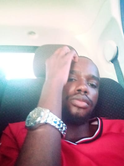 The image depicts an adult male seated within the interior of a vehicle in Majengo ward, Kenya. The individual has dark skin, short dark hair, and a neatly trimmed beard. He is wearing a red short-sleeved shirt with a black and white striped collar. His right arm is raised, with his hand resting on his forehead or hairline. A prominent silver-toned wristwatch with a patterned band is visible on his left wrist. The man is looking directly at the camera with a neutral expression. The vehicle interior shows a light-colored headliner and a dark headrest behind the subject, along with a portion of a light-colored vehicle pillar on the right side. Bright ambient light originates from the left side of the frame.
