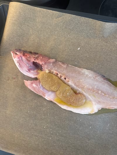 An overhead view displays a raw, gutted, and butterflied fish, oriented with its head to the left, resting on a sheet of light brown parchment paper. The fish's internal cavity contains two yellowish-tan roe sacs. Coarse salt crystals are scattered over the pinkish-white fish flesh and the roe. Yellowish oil is visibly pooled beneath and around the roe. The parchment paper lies on a dark, flat surface, partially visible at the top edge of the frame, suggestive of a baking tray or stovetop. The scene depicts a culinary preparation step for the fish, likely prior to cooking. No individuals are present. The location, Ericeira, Portugal, is provided as contextual information and is not visually discernable within the image itself.