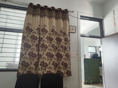 The image shows an indoor scene, likely a room in Dhandhuka, India. In the foreground, two dark-colored chairs are partially visible. A patterned curtain, with a beige background and floral motifs in brown and darker beige, hangs over a window that has horizontal bars. To the right of the curtain, a square clock with Roman numerals is mounted on a light-colored wall. Further to the right, a doorway leads to another area where a dark green metal cabinet is visible. A dark backpack rests on top of the cabinet. The overall environment appears to be an office or a similar functional space.