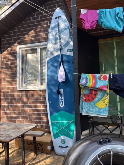 In the city of Kzyl-Bairak, Russia, a blue and white paddleboard stands upright against a brick building. A paddle is attached to the paddleboard. Nearby, on a clothesline, hang pink and blue swim bottoms and a colorful towel with fruit designs. To the left, a wooden table sits on sand. To the right, a foldable chair is placed next to the paddleboard. Partially visible in the foreground is a dark inflatable ring. The scene is outdoors, with sunlight casting shadows.
