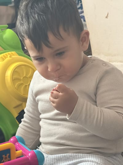 A close-up, medium shot captures a young child, possibly a toddler, with dark hair and fair skin. The child is wearing a light-colored, long-sleeved shirt and light gray pants. The child is looking downwards, with their mouth slightly open, and their right hand is held up to their face, near their mouth. The child appears to be eating or holding something small and red. In the background, to the left, a large, bright yellow and green plastic toy is visible. The background wall is a plain, light color. The lighting suggests an indoor setting. There is no discernible information to place this image within Mid Devon, United Kingdom.