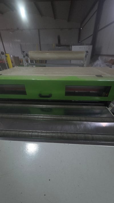 The image shows a section of what appears to be an industrial setting. Visible is a large piece of machinery, partially covered in protective wrapping. It has a green cabinet-like structure with a handle, alongside vertical metal panels. To the left, further in the background, there is a tall, cylindrical object also wrapped, suggesting it's a component of the machinery. The environment is enclosed, possibly a warehouse or factory, with a corrugated metal roof visible in the upper left corner of the image. The overall impression is an indoor, potentially manufacturing-related space. The location in the city of Balgarevo, Bulgaria is not discernible from the image.