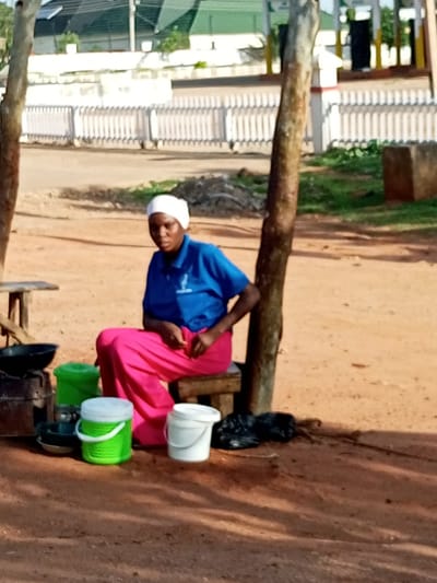 A woman is seated on a low wooden stool next to a cooking apparatus and several containers on a dirt ground. She is wearing a white headscarf, a blue t-shirt with a small logo, and bright pink trousers. To her left, a dark pan sits atop what appears to be a charcoal stove. In front of the stove are two plastic buckets, one green and one white, both with lids. Behind the woman, a rough-hewn wooden post is visible, and in the background, a white picket fence and buildings are discernible. The scene appears to be an outdoor market stall in Wuro Dole, Nigeria.