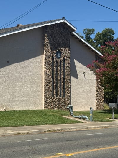 The image shows the side of a building with a stucco exterior and a stone facade accenting the corner where a set of three tall, narrow windows are located. A dove symbol is visible in the upper portion of the windows. The building has a sloped roof with dark shingles and white trim. To the right of the building, a tree with pink flowers is visible, as are two mailboxes. In the foreground, there is a grassy area bordering a sidewalk, and a street with a yellow line painted on it. A set of pipes and valves are visible on the grass near the sidewalk. The sky is clear and blue. This scene is located in Rio Linda, United States of America.