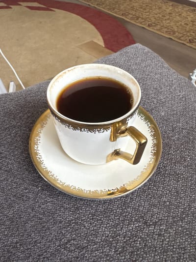 A close-up, overhead shot shows a white teacup with gold trim and a gold handle filled with dark coffee, resting on a matching saucer. The cup and saucer are placed on a dark gray, textured surface, possibly a sofa or cushion. In the blurred background, a rug with a circular red and tan pattern is visible. A white cable or string runs from the upper left corner into the frame. The scene is indoors and does not provide specific visual cues to pinpoint a location in Таганрогский, Russia.