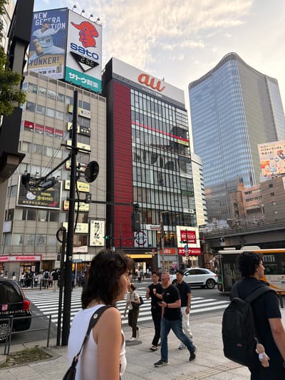 The image depicts a street scene in Chiyoda, Japan, likely during daylight hours given the overcast sky. The foreground features a woman in a white tank top and glasses, with her back to the camera, carrying a dark shoulder bag. To her right, a man with a black backpack walks away from the camera. In the mid-ground, several pedestrians are crossing a zebra crossing, including a man with a beard wearing a black t-shirt and jeans, looking at his phone, and two other men in black. A white SUV is also visible on the road.

The background is dominated by tall buildings with numerous signs. One prominent building displays an "au" logo, suggesting a telecommunications company. Another large billboard features a baseball player and the word "YUNKER!". Across the street, a modern skyscraper with a curved glass facade rises high into the sky. An elevated pedestrian bridge or railway line is visible in the distance. The overall environment is urban, with a mix of modern architecture and advertising.