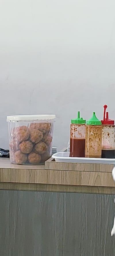 A clear plastic tub filled with meatballs sits on a wooden counter to the left of the image. Next to it, on a white tray, are three squeeze bottles of sauces. The first bottle, on the left, contains a dark red sauce. The middle bottle has a light brown sauce, and the third bottle, on the right, contains a dark brown liquid. The background is a plain white wall. The scene appears to be a food stall or a restaurant in Medan, Indonesia.