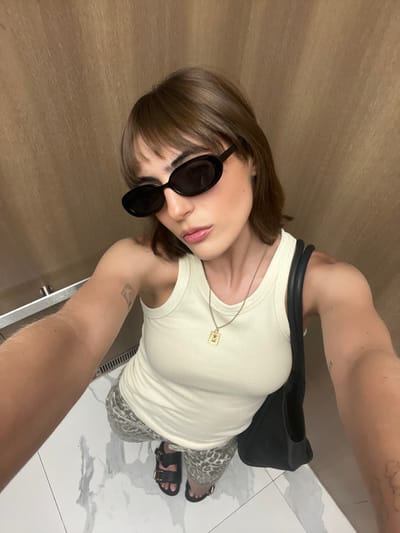 A female subject is depicted in a high-angle selfie, taken within what appears to be an elevator or a small room. The location is Sunny Isles Beach, United States. The subject has light brown hair styled with bangs and a bob cut. She wears black oval sunglasses, an off-white ribbed tank top, and light-colored patterned pants, possibly an animal print. Her footwear consists of black two-strap sandals with gold buckles. A gold-toned necklace with a rectangular pendant featuring an embossed design is visible around her neck. She carries a black shoulder bag on her left side. A light-toned tattoo is present on her right upper arm. The background consists of uniform light brown, vertically textured walls, and a white floor with grey marble-like veining. A silver metallic bar or handrail and a horizontal ventilation grate are visible on the left side of the frame.