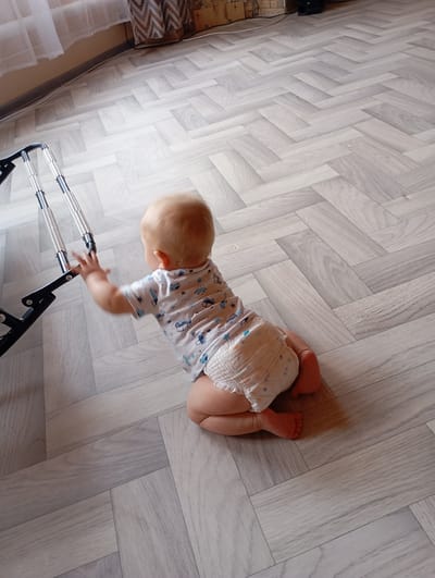 A baby wearing a diaper and a light-colored shirt with a pattern of cars and stars is crawling on a light-colored floor with a herringbone pattern. The baby is reaching out with its left hand towards a black walker with silver springs. The background shows a curtain and a wall. The location is not specified as being in the city of Vysokaya Gora, Russia.