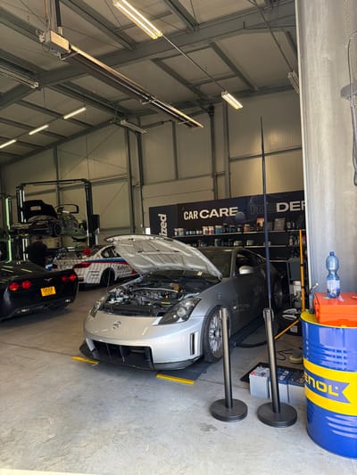 This image depicts the interior of an automotive service facility located in Meuspath, Germany. The scene is illuminated by overhead fluorescent lighting and ceiling-mounted strip heaters, revealing a concrete floor and white, corrugated metal walls with exposed metal ceiling trusses.
In the foreground, a silver Nissan 350Z sports car is positioned with its hood open, exposing the engine bay. The vehicle features a modified front splitter and aftermarket wheels, resting on yellow diagnostic pads. Two black stanchions stand vertically to the right of the Nissan. Adjacent to these, a blue and yellow industrial drum is visible, topped with an orange first-aid kit and a clear plastic water bottle. Two car batteries are on the floor near the drum.
Further into the facility, a black Chevrolet Corvette (C6 generation) is parked with its rear facing the viewer, displaying a German license plate "GK55 CGO". Behind the Corvette, a white BMW M-series car with a multi-colored racing livery, including "Safety first" text, has its trunk open. In the background, to the left, a dark-colored sports car is elevated on a hydraulic lift, also with its hood open, suggesting ongoing service. One male individual, dressed in dark clothing, is partially visible near the rear of the Corvette, facing towards the BMW.
Along the far wall, a prominent black sign reads "CAR CARE RIEDER," with shelves beneath displaying various automotive products and detailing supplies.