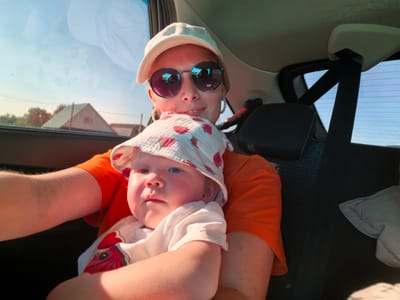 In the city of Karaiganovo, Russia, a woman and a baby are in a car. The woman, wearing an orange t-shirt, a white baseball cap, and round sunglasses, holds the baby. The baby is wearing a white shirt with a red graphic and a bucket hat with a strawberry pattern. They are both positioned in the front passenger seat of the vehicle. Through the car's windows, a clear sky and some houses are visible, suggesting an outdoor setting during daylight. The interior of the car is partially visible, with a black headrest and a seatbelt mechanism evident.
