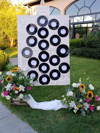 This image depicts an outdoor scene, likely at a wedding reception, on a grassy lawn in Scandiano, Italy. A large board covered in sheet music serves as a backdrop, adorned with vinyl records arranged in a grid pattern. Each record has a label indicating a song title. In the foreground, a floral arrangement of sunflowers, roses, lilies, and other assorted flowers rests on a small wooden stand draped with white fabric. Behind the floral display and to the right, a man in a light brown jacket is visible walking towards an entrance. The setting appears to be during daylight, with dappled sunlight illuminating the scene. The architecture visible in the background suggests a building with large arched windows, characteristic of Italian villas or event venues.