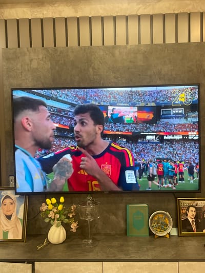The image displays a living room setting with a large television screen dominating the scene, showing a football match. On the screen, two male footballers are engaged in an intense conversation on the field, with a stadium full of spectators in the background. The left player wears an Argentina jersey, while the right player is in a red and yellow Spanish national team kit. Above the players, the ITV logo and a "20" are visible. The television is mounted on a textured grey wall.
In front of the television, on a grey surface, are several decorative items. To the left, a framed portrait of a woman in a hijab is positioned. Next to it, a white vase holds yellow tulips and delicate pink flowers. To the right of the vase is a clear glass cloche with a lid. Further to the right, a small green book and a decorative plate with an embossed image are placed side-by-side. A framed portrait of a man in a suit is visible on the far right. The overall environment suggests a domestic space. The presence of the ITV logo and the Spanish national team suggests a broadcast of a football match, likely during the FIFA World Cup, given the stadium context and the "FIFA WORLD CUP" text visible on the screen. The items on the shelf, including the religious book and portraits, indicate personal belongings and cultural context. There is no specific visual information within the image that directly places the scene in Sumgayit, Azerbaijan.