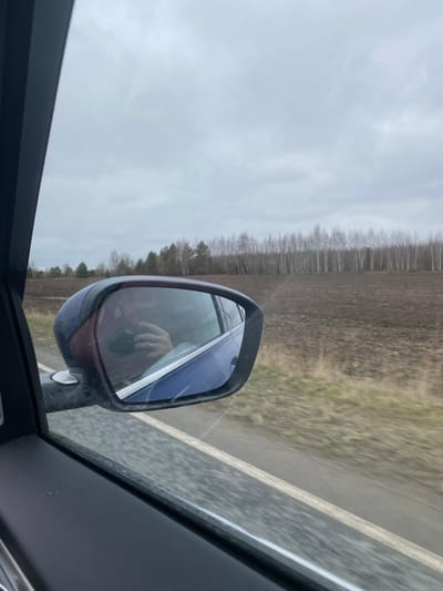 The image shows a view from inside a moving car, with the side mirror visible in the foreground. The reflection in the side mirror shows the face of a person, likely the driver, and their hand holding a device, possibly a phone. The background seen through the car window and in the side mirror shows a rural landscape with a field, trees, and a cloudy sky. The location is likely Doksha, Russia. A paved road is also visible alongside the field.