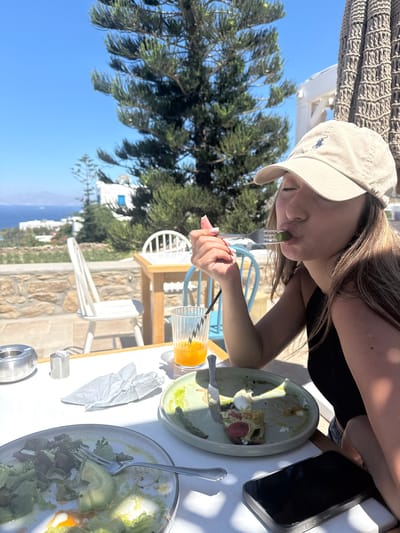 A young woman wearing a beige baseball cap and a black top is eating on a fork in an outdoor cafe in Mykonos, Greece. She is sitting at a white table with food and drinks. In the background, there is a view of the sea and traditional white-washed buildings. The weather is sunny and clear.