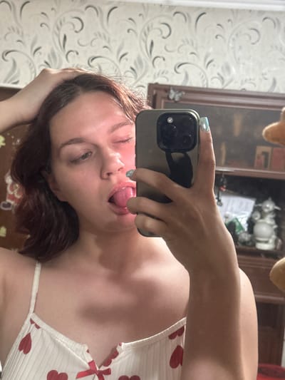 A young woman is taking a selfie in a mirror. She is wearing a white camisole with red hearts. She is sticking her tongue out and winking. The background is a bedroom with floral wallpaper and a wooden dresser. The image does not contain enough information to determine the specific location in Bishkek, Kyrgyzstan.