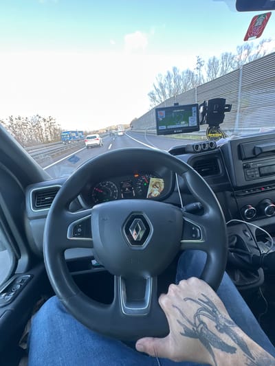 The image depicts the interior of a vehicle, likely a commercial van, from the driver's perspective, traveling on a multi-lane highway under clear daylight conditions.

The vehicle is located on the A8 Innkreis Autobahn in Austria, in the vicinity of Kematen am Innbach, heading east towards Wels and Linz as indicated by the navigation system.

**Interior Scene:**
A driver's left hand, adorned with a large black and grey tattoo of two bird-like figures with outstretched wings, rests on the bottom spoke of a black, multi-function steering wheel with a central silver Renault emblem. The driver's legs, clad in blue jeans, are visible below the steering wheel. The instrument cluster behind the steering wheel displays a speedometer indicating approximately 120 km/h and a tachometer around 2000 RPM. A religious icon is tucked into the right side of the instrument cluster. A black aftermarket GPS device (VIVAVAS) is mounted on the windshield, displaying a map of the A8 highway and showing "A8 WELS | A1 LINZ" with "28km 21:00 Wien". Adjacent to the GPS is a black smartphone holder, and an "Areon" brand air freshener hangs from the rearview mirror area. A strip of three 12V power outlets is visible on the dashboard below the GPS. A red and white sticker is affixed to the upper right corner of the windshield.

**Exterior Scene:**
The vehicle is on a paved highway with at least two lanes for the direction of travel. Moderate traffic is present, including a white passenger car in the left lane and a blue truck with two trailers in the right lane ahead of the vehicle. A tall, grey sound barrier wall lines the right side of the highway. Beyond the barrier and on the left, leafless deciduous trees are visible. The sky is light blue with sparse white clouds.