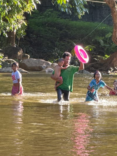 In Ranong, Thailand, a group of children and a man are wading in a shallow, muddy river. Sunlight filters through the dense foliage lining the riverbank, creating dappled patterns on the water's surface. A man in a green shirt carries a young child in his arms and holds a pink inflatable ring aloft. To his left, a young girl in a white shirt and pink skirt stands facing the camera. To the right, a girl in a blue and green patterned dress splashes in the water, with another child partially visible in the background. The riverbed appears to be composed of sand and rocks. The overall impression is one of a casual, recreational outing in a natural, tropical setting.