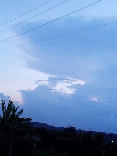 The image depicts an outdoor scene within Nachu ward, Kenya, during what appears to be twilight. The upper portion of the frame features a blue sky with a mixture of dense grey and lighter white clouds. Three dark, parallel power lines are visible spanning horizontally across the upper left quadrant. The left foreground is occupied by the dark silhouette of large-leafed vegetation, consistent with banana plants. The midground and background reveal a dark, undulating horizon line, indicative of distant hills or dense tree cover, with faint, indistinct structural elements intermittently visible beneath the horizon. No individuals or specific activities are depicted.
