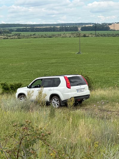 In the village of Humentsi, Ukraine, a white Nissan X-Trail SUV is parked on the side of a grassy field. The SUV is positioned facing right, with its rear to the viewer, revealing its license plate, which reads "BX 8662 IC." The vehicle is situated amidst tall, dry grass and scattered wildflowers, with a lush green field stretching out behind it. In the distance, a line of trees borders the horizon, beyond which are rolling hills and a cloudy sky. The scene appears peaceful and rural.