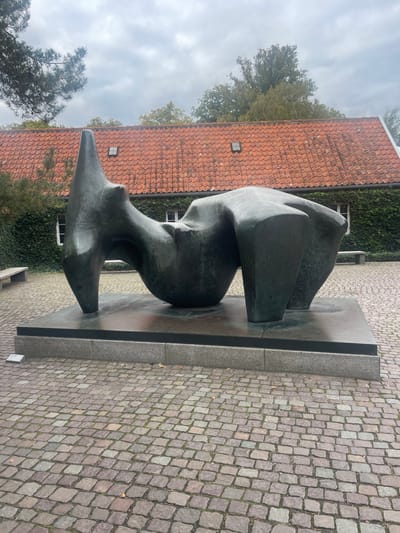 A large, patinated bronze sculpture, "Reclining Figure No. 5" by Henry Moore, is prominently displayed outdoors on a cobbled plaza in Fredensborg, Denmark. The abstract form, characterized by fluid, organic shapes, rests on a dark, polished rectangular slab, which is supported by a wider, lighter gray granite plinth. The ground is paved with variegated grey and reddish-brown cobblestones arranged in an interlocking pattern. In the background, a building with a distinct red tiled roof and walls densely covered in green ivy is visible. Multiple white-framed windows are discernible through the ivy. Overcast grey skies are above, with green deciduous trees extending beyond the roofline. A pine branch with needles is visible in the upper left corner. Two light-colored benches are positioned against the ivy-clad building wall in the mid-ground. A small white rectangular plaque is visible on the cobblestones near the sculpture's base on the left. No individuals are present.