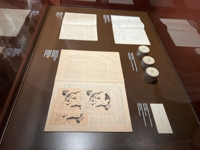 The image shows an exhibit in the Trakai History Museum in Trakai, Lithuania. The exhibit displays historical documents and artifacts. In the foreground, a large open ledger book is visible with handwritten notes and a series of hand-drawn illustrations depicting maps or diagrams. To the right of the ledger, three cylindrical objects, possibly film canisters, are arranged in a row, each with a label indicating a year, such as "1937" and "1938." Behind these objects, an open book with handwritten text and diagrams is displayed. Further to the left, another open book with handwritten text is visible, partially obscured. The exhibit is presented on a dark brown surface under display lighting, suggesting an indoor museum setting. A label with text in Polish and English, including "Walery Romańczuka's Business Card," is placed to the lower right of the ledger.