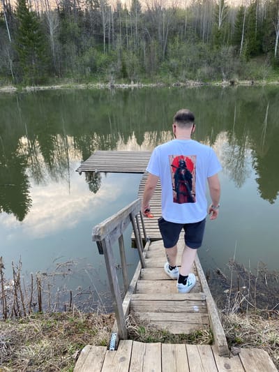 A man stands on a wooden dock extending into a body of water. He faces away from the camera, wearing a white T-shirt with a large graphic on the back, dark shorts, and white sneakers with black socks. The dock has a small wooden ladder leading down into the water. The water reflects the trees lining the opposite bank. The location is Баклуши, Russia.