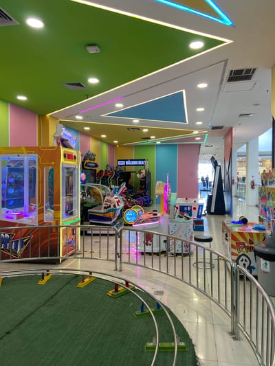 The image shows an indoor arcade in Kudus, Indonesia. The arcade features a variety of colorful game machines, including claw machines, racing games, and air hockey tables. The ceiling is painted in vibrant colors with neon lighting, creating a lively and energetic atmosphere. In the foreground, a small train track with green turf is visible, suggesting a children's ride. A metal railing encloses the track and separates it from the rest of the arcade. Several game machines are positioned behind the railing, and some people can be seen in the background, interacting with the machines or walking through the arcade. The overall impression is of a fun and engaging entertainment venue.