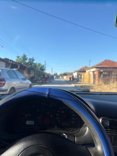 The image is taken from the interior of a car looking out through the windshield on a sunny day. The dashboard and steering wheel are visible in the foreground, with the steering wheel covered in a blue protective sleeve. The road ahead leads through a village with houses on either side. The houses have red tiled roofs and appear to be of simple construction. Trees and dry vegetation are visible along the roadside. The sky is clear blue. The car's odometer displays "248". The scene depicts a typical day in a Bulgarian village, potentially in the city of Vinitsa, Bulgaria, given the context.