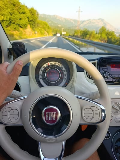 A first-person perspective shot from inside a white Fiat 500 shows a driver's hand on the steering wheel, driving on a road in Stanišići, Montenegro. The dashboard features a circular speedometer with a digital display showing the date "29 Jun 19:33" and the odometer reading "90974 km." The car's radio is on, displaying "PLAY" and a website address. The road is flanked by lush green trees and mountains in the background. The sky is clear and blue.