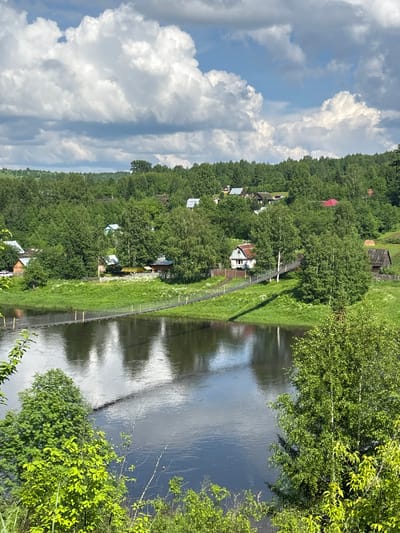 The image depicts a serene riverside landscape in Gubakha, Russia. A suspension bridge, constructed with dark planks and supported by cables, spans a calm, dark river. The river's surface reflects the overcast sky and the surrounding greenery. On the opposite bank, a verdant hill is densely covered with lush green trees, interspersed with several small, traditional wooden houses, some with red or grey roofs, and a few scattered structures like sheds or barns. The architecture suggests a rural or semi-rural setting. The foreground features vibrant green foliage and trees, framing the view of the river and the village. The sky above is a mix of blue and large, white cumulus clouds, indicating fair weather. The scene conveys a sense of tranquility and natural beauty.