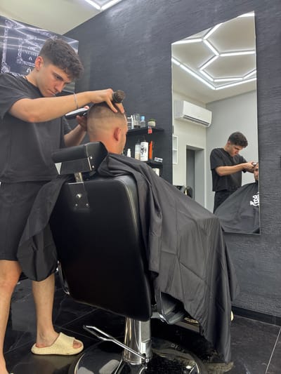 This indoor photograph, taken in Simitli, Bulgaria, captures a barbershop scene with a modern, dark aesthetic. The image is oriented sideways, with the main subjects appearing rotated.
In the foreground, a young male barber with dark, curly hair, wearing a black t-shirt and shorts, is intently working on a client's hair. He wears a silver chain bracelet on his left wrist and holds a hair brush near the client's head. The client, positioned in a black upholstered barber chair and draped with a black cape, has very short, buzzed hair and is looking upwards. His light-colored, slip-on sandals, adorned with a repeating text pattern, and lower legs are visible beneath the cape on the dark, possibly tiled floor.
A large rectangular mirror dominates the background, reflecting the barber from a different angle as he works. The reflection also shows parts of the barbershop's ceiling, featuring a striking geometric pattern of bright, hexagonal light fixtures, and a white air conditioning unit mounted on the wall. Behind the barber chair, shelves stocked with various hair care products are partially visible. A poster with the word "UNLEASHED" and an ornate design is also faintly visible in the reflection.
The atmosphere appears focused and professional, with both individuals engaged in the haircutting process. The bright artificial lighting suggests either daytime or a well-lit interior. The predominant colors are black, white, and grey, contributing to the contemporary feel of the establishment.