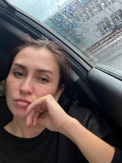 This media file is a selfie taken by a person in a car. The subject is a young woman with brown hair and freckles on her face. She is wearing a black top and is resting her hand on her chin. Her gaze is directed towards the camera, conveying a contemplative or relaxed emotion.
The setting appears to be inside a vehicle, indicated by the interior of the car visible in the foreground. Through the car window, a building with multiple windows is visible, and the glass is covered in raindrops, suggesting it is raining. The building appears to be a residential or office building.
The time of day is not explicitly indicated but the lighting suggests it could be daytime. The weather is clearly rainy. The location context provided is Verkhneyarkeyevo, Russia, which adds a geographical element to the scene. There is no visible text in the image.