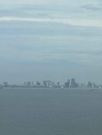 This is a wide-angle, aerial shot of a sprawling city skyline situated across a body of water. The city, identified as Hải Vân Ward, Vietnam, features a dense collection of high-rise buildings, including skyscrapers, apartment complexes, and what appear to be under-construction towers. The buildings are varied in height and architectural style, with some being modern glass structures and others more traditional.
The body of water in the foreground is calm and appears to be a bay or a harbor, with several vessels visible. These include cargo ships, smaller boats, and potentially ferries, dotting the surface of the water.
The sky above is overcast, with layers of clouds creating a muted, diffused light that casts a soft glow over the scene. The overall atmosphere is serene, with the city lights subtly visible in the distance, suggesting it is either early morning or late afternoon. The horizon line is sharp, separating the cityscape from the expanse of water and sky. There are no visible people in the frame, and the focus is on the urban landscape and its relationship with the surrounding environment.