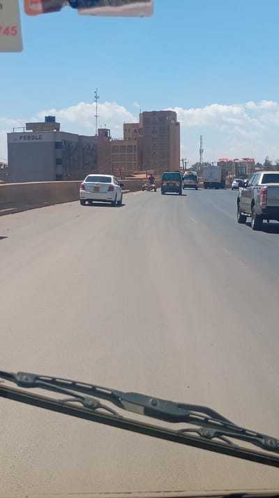 Vehicles of various types travel on a wide, paved road under a bright sky. A white sedan, a van, a small cart pulled by a person, a delivery truck, and a pickup truck are visible. Buildings, including one labeled "PEBBLE," rise in the background, along with a communication tower. The scene is captured from the perspective of a vehicle, as indicated by the windshield wiper in the foreground.