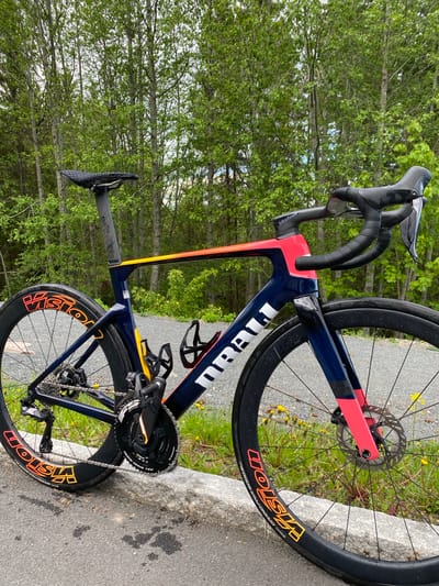 The image depicts a high-end road bicycle resting on a gray concrete curb. The bike is predominantly dark blue with red and yellow accents. It features a carbon fiber frame, disc brakes, and Vision wheels with Continental Grand Prix 5000 tires. The bicycle's components include black handlebar tape, a water bottle cage, and a black seat.

The background is a natural setting, featuring a dense forest with green trees. The scene appears to be taken on a paved path or road near the forest. The location is identified as being in Lommedalen, Norway. There are no people visible in the frame. The photo appears to have been taken outdoors during daylight hours.