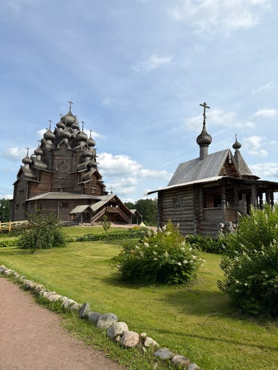 This is a full-frame, eye-level, outdoor shot of two wooden churches in Nevskii Parkleskhoz, Russia. The larger church on the left is tall and features numerous onion domes, each topped with a cross. The smaller church on the right is lower and wider, also with onion domes and crosses. Both structures are made of logs and exhibit traditional Russian wooden architecture.

The churches are set within a green, grassy park. A gravel path runs along the bottom left of the image, bordered by a line of large, gray stones. In the foreground, there are several bushes with white flowers. The background consists of green trees and a partly cloudy blue sky. No people are visible in the image. The lighting suggests it is daytime, with bright sunlight casting shadows.