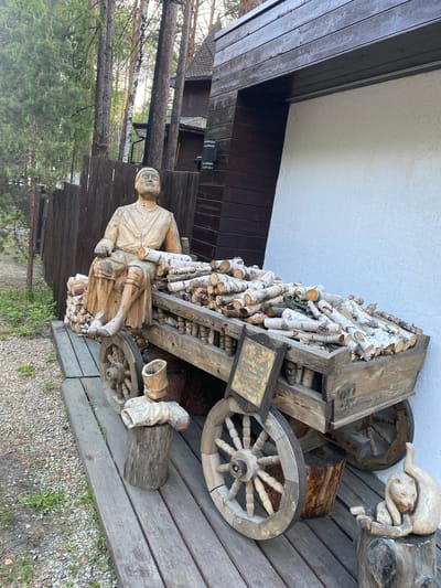 The image depicts an outdoor scene in Tal'tsy, Russia, featuring a wooden sculpture of a man sitting on a cart filled with logs. The cart has wooden wheels and is made of rough-hewn planks. The man sculpture is carved with detailed clothing and facial features, and a few logs are placed beside him as if he is holding them. Other wooden carvings, including a cat sculpture, are visible on the wooden platform where the cart is situated. The background includes a wooden fence, tall trees, and the exterior of a building with dark wooden siding and a white plastered section. The ground is covered with gravel and some vegetation. The scene appears to be part of an outdoor exhibition or decorative display.