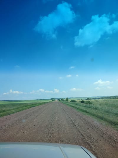 The image depicts a gravel road stretching into the distance, bordered by vast, green fields under a bright blue sky with scattered clouds. The perspective suggests the photo was taken from a vehicle, with the hood visible in the foreground. The landscape appears rural and open. There is no visible indication of the city of Bakhmut, Russia, nor are there any people or specific identifiable objects other than the natural environment and a vehicle.
