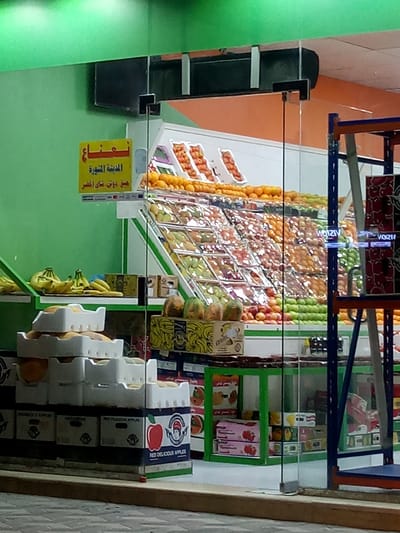 The image shows a view through the glass storefront of a fruit and vegetable market, likely located in Al Khobar, Saudi Arabia. Inside the store, a multi-tiered display case presents a variety of fruits, including oranges, apples (both green and red), and other produce. Boxes of "Red Delicious Apples" are stacked near the entrance. A yellow sign with Arabic script is visible above the fruit display, possibly indicating the name of the market or a special offer. On the left, there are bananas and melons. A blue metal shelving unit is positioned toward the right side of the frame. The store's lighting and color scheme consist of green walls, an orange ceiling, and the vibrant colors of the fruits on display. The street outside appears to be deserted.