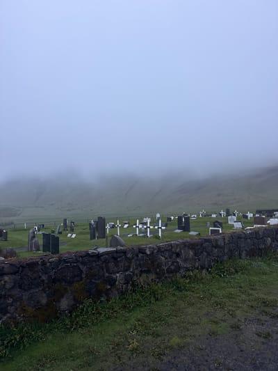 This image depicts a cemetery in Vik, Iceland, on a foggy day. The landscape is characterized by a grassy field that slopes upward into the fog. Scattered across the field are numerous headstones and crosses, indicating grave markers. A dark stone wall runs along the right edge of the frame, separating the cemetery from a path or road covered in grass and gravel. The fog obscures the distant features, creating a somber, ethereal atmosphere. There are no people visible in the frame.