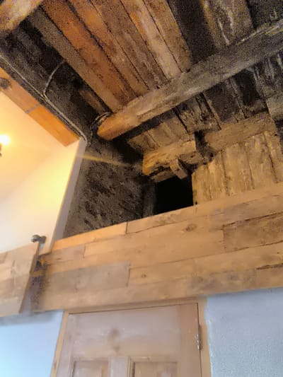 The image shows a dark, narrow opening between wooden beams and stone walls, suggestive of an attic or crawl space. A light fixture is visible to the left, illuminating part of a white wall. The overall impression is one of aged construction and a utilitarian space. The location in Kuldīga, Latvia, is not visually discernible from the image itself; it is an external piece of information.