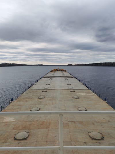 The image shows a view from a ship's deck on a body of water, likely a river or a wide lake, under a cloudy sky. The deck is tan and marked with several round, bolted hatches. A white railing is visible in the foreground. Across the water, forested land is visible. The scene is taken in Khoroshevo, Russia.