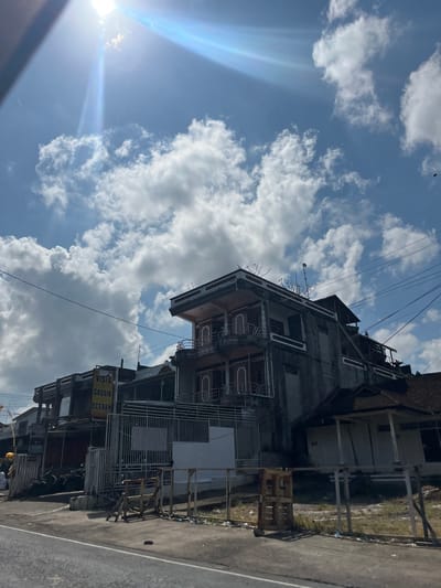 The image shows a building with a sign that reads "VISTA GROSIR ECERAN". The building appears to be under construction or in disrepair, with unfinished concrete walls and exposed rebar. There are several floors with balconies and windows. A street runs in front of the building, with a sidewalk and some debris on the ground. The sky is blue with scattered white clouds, and the sun is shining brightly, suggesting it is daytime. The scene appears to be in Kerambitan, Indonesia. The overall impression is one of urban development or decline, with a mix of modern architecture and signs of neglect.