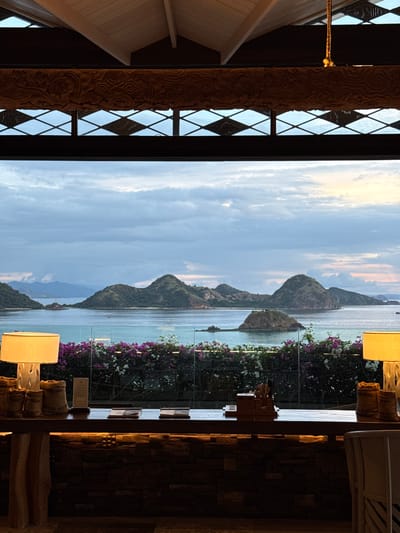 The image presents a view of islands and water through a window, likely from an establishment in Labuan Bajo, Indonesia. The seascape features a series of mountainous islands stretching into the distance, beneath a partly cloudy sky. In the foreground, white and purple flowers adorn the border of what appears to be a glass partition. Adjacent to the floral display, elements of interior design, including lamps and textured walls, are partially visible, hinting at a seating or waiting area within a building. On the left side of the image, an ornate, carved wooden structure with diamond-patterned windows frames the view, adding to the architectural context of the scene.